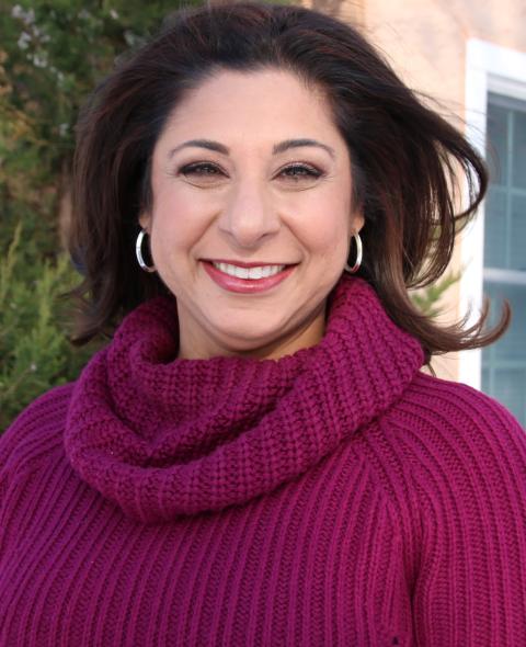 A feminine-presenting person with medium-light skin tone and brown eyes smiles into the camera. They are wearing a thick magenta turtleneck and medium-sized hoop earrings. They stand in front of a the corner house with windows on both sides. A tall evergreen tree slants into the house directly behind them.