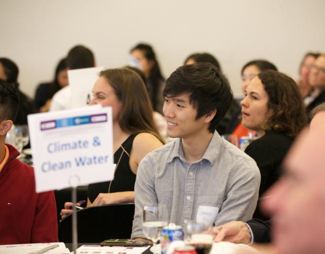 Student attends SDG Climate Event Young man in focus sitting at a table with a sign reading Climate & Clean Water.