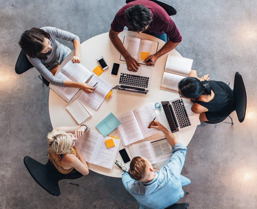 aerial view of five people sitting at a round table, with laptops, books, and notes strewn about
