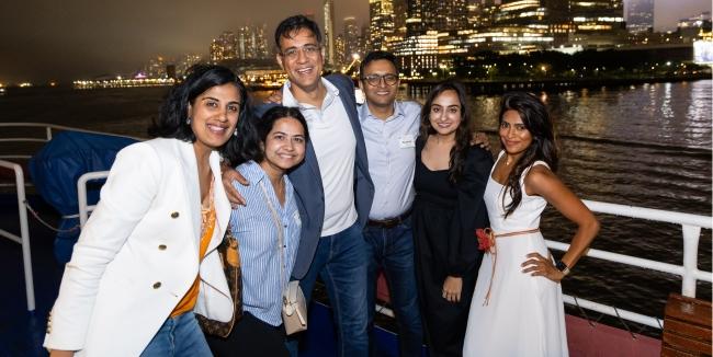 A group of 6 people stand outside at night on the deck of a boat with the NYC skyline in the background