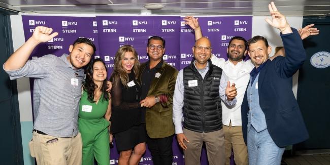 A group of 7 smiling people standing in front of a step-and-repeat that says "NYU Stern"